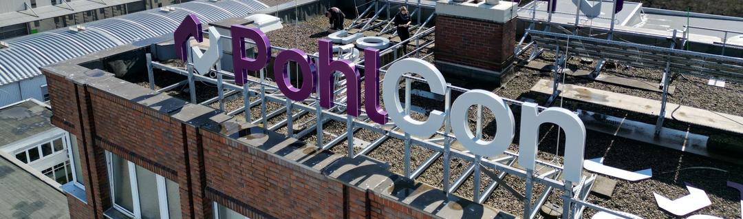 Drohnenaufnahme des PohlCon-Hauptgebäudes mit großer Dachwerbung in Montage, Blick über Gewerbedächer und grüne Landschaft.