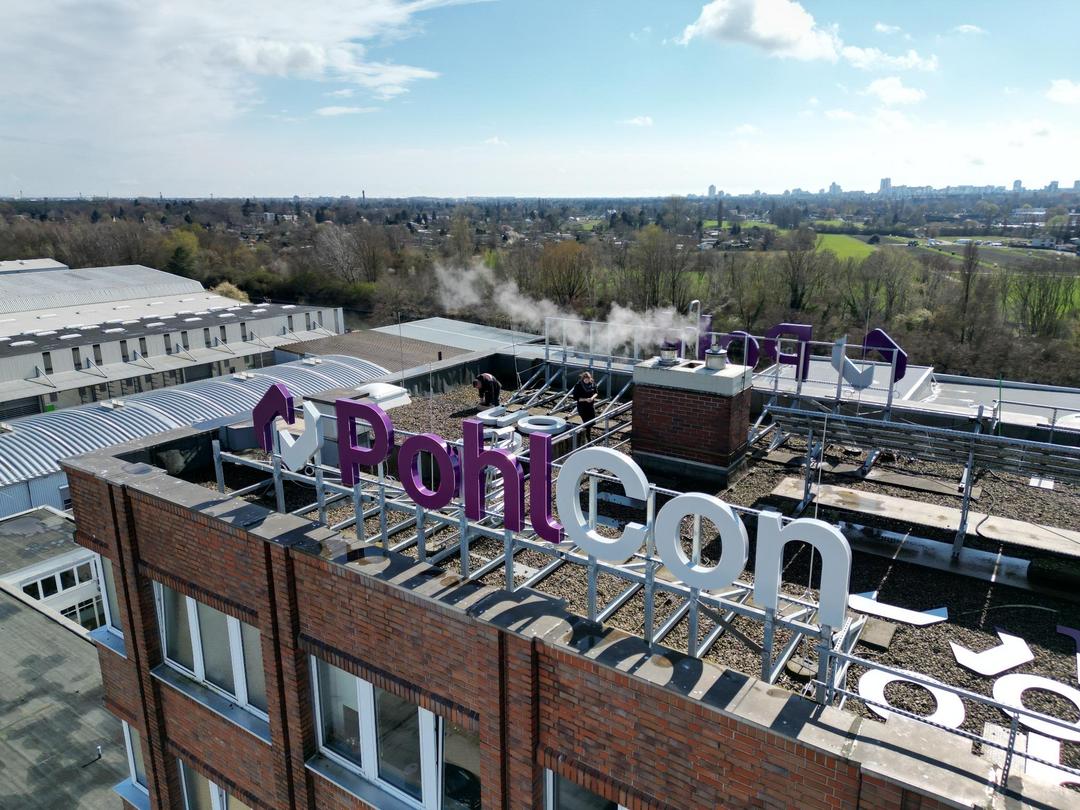 Drohnenaufnahme des PohlCon-Hauptgebäudes mit großer Dachwerbung in Montage, Blick über Gewerbedächer und grüne Landschaft.