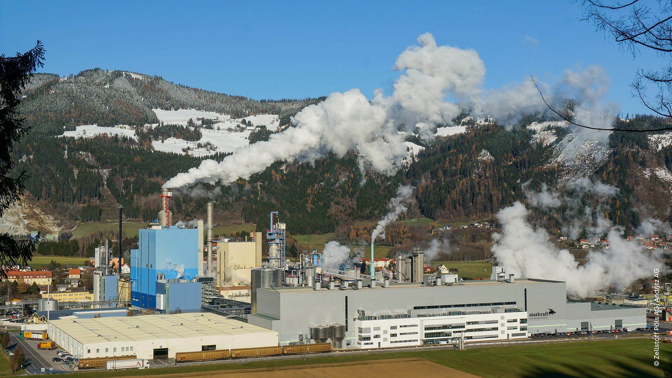 Industriegebäude mit aufsteigendem Rauch aus Schornsteinen, im Hintergrund schneebedeckte Gebirgskette und blauer Himmel.
