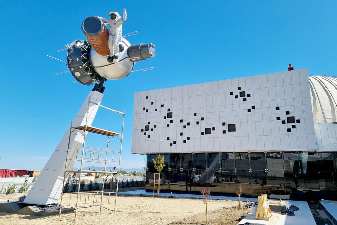 Skulptur eines Astronauten auf einem Raumschiff vor modernem Gebäude und blauem Himmel