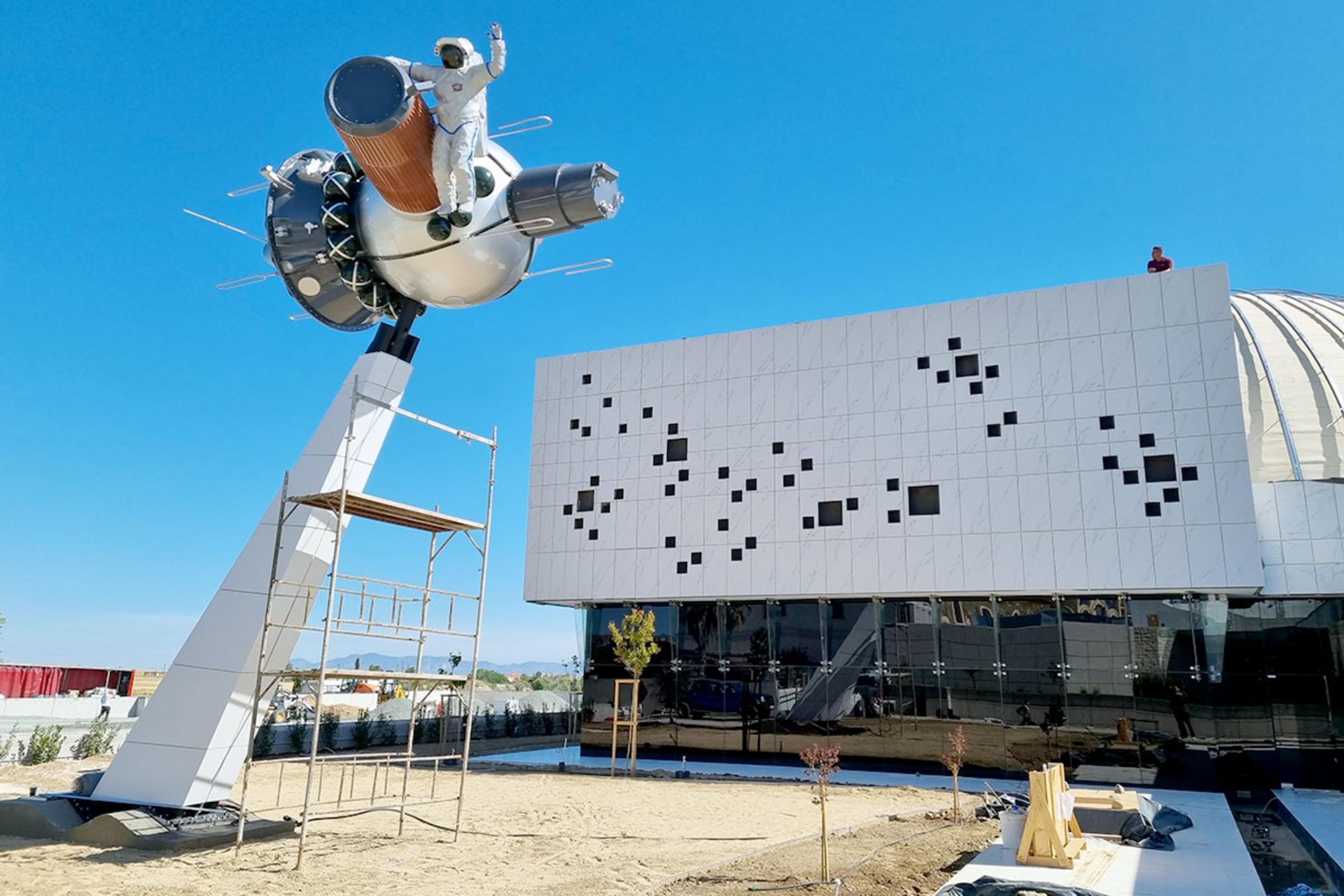 Skulptur eines Astronauten auf einem Raumschiff vor modernem Gebäude und blauem Himmel