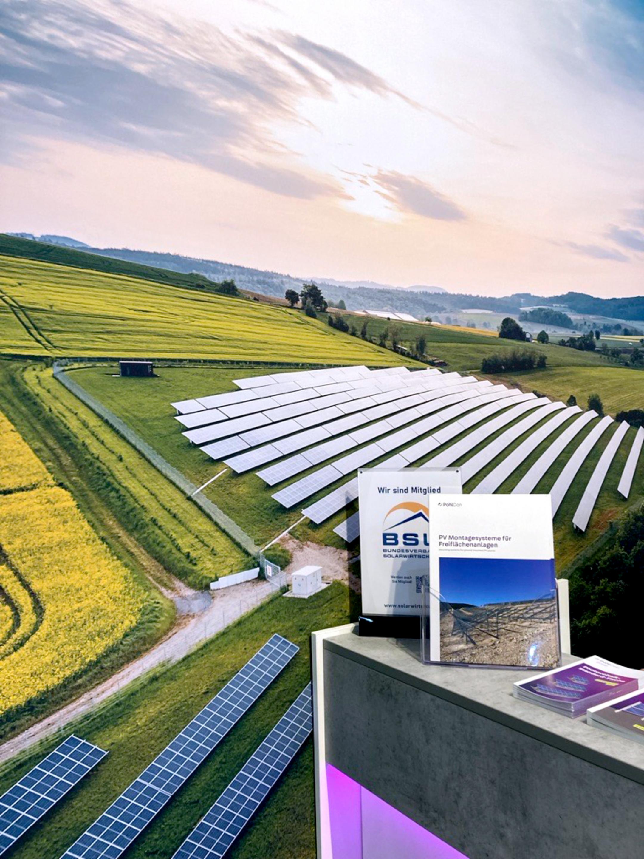 Messetresen mit Broschüren darauf vor einer großen Fotowand, die eine Photovoltaik-Freiflächenanlage zeigt.