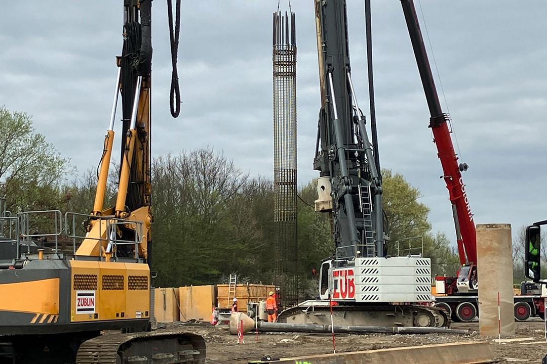 Baustelle mit mehreren Großgeräten beim Einheben eines Bewehrungskorbs; Arbeiter in Warnkleidung bei bewölktem Himmel