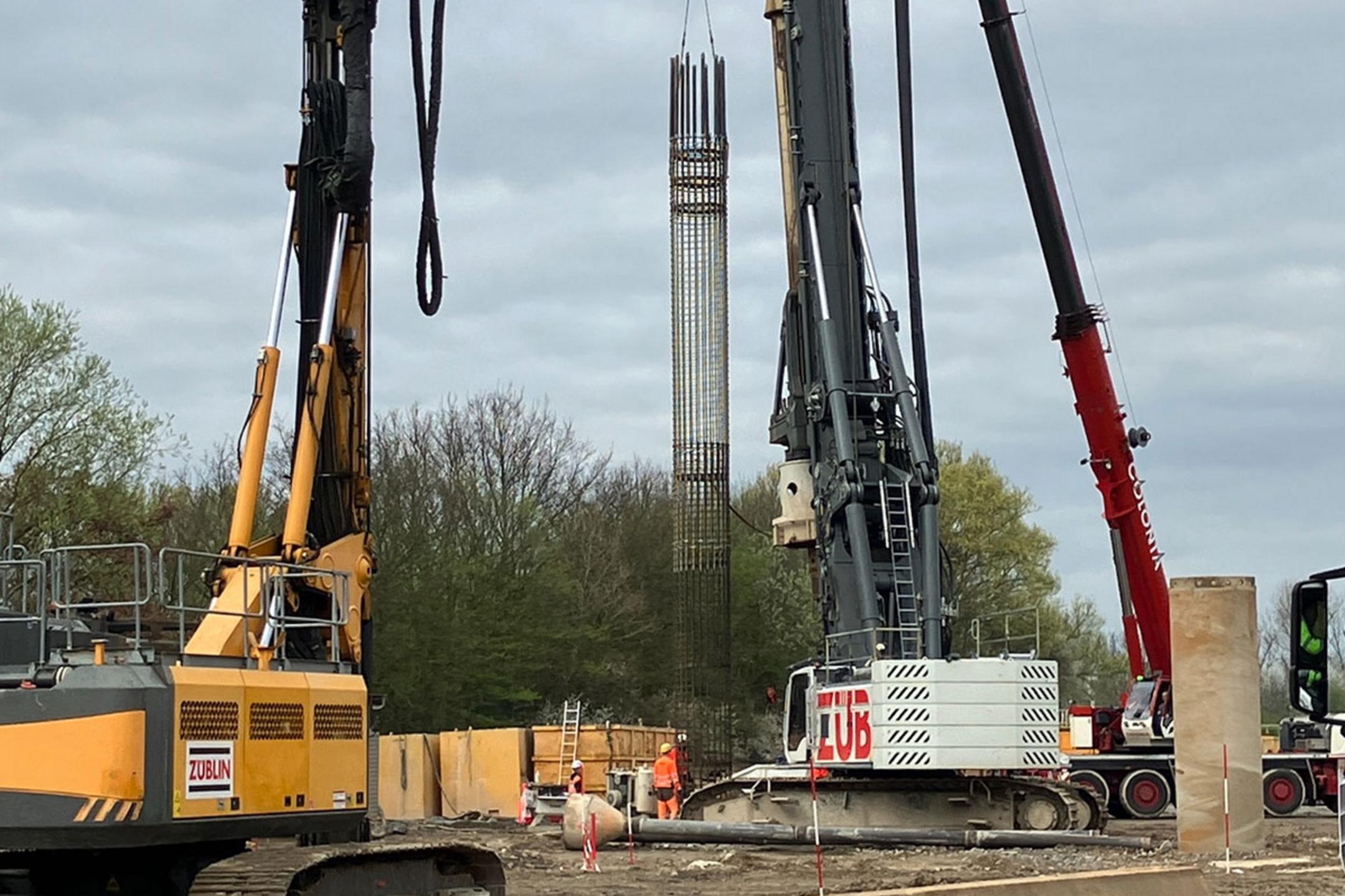 Baustelle mit mehreren Großgeräten beim Einheben eines Bewehrungskorbs; Arbeiter in Warnkleidung bei bewölktem Himmel