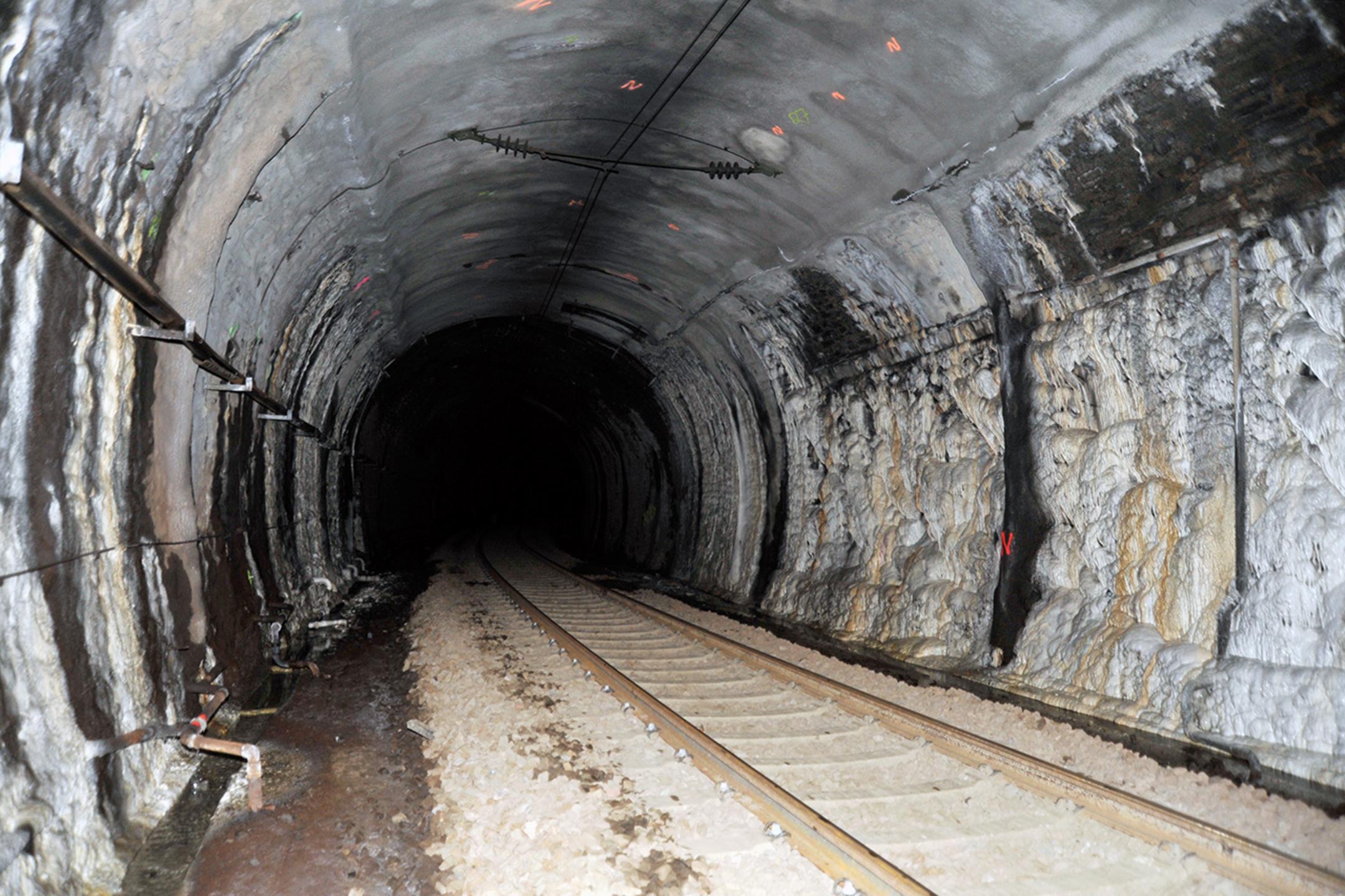 Dunkler Eisenbahntunnel mit markanten Kalkablagerungen an den Wänden, Schienen