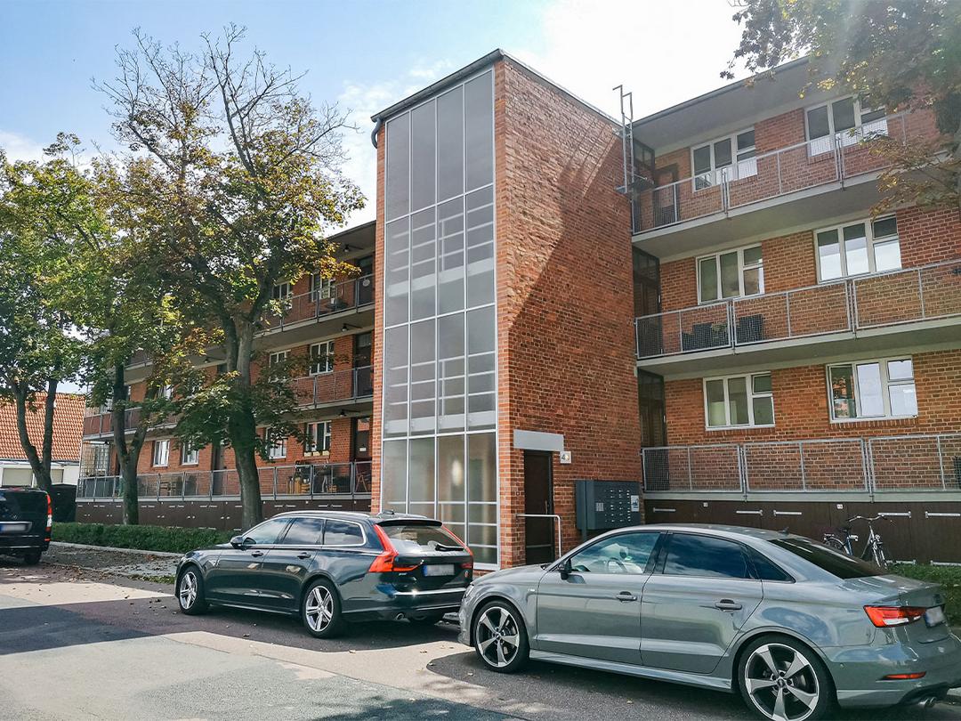 Mehrfamilienhaus mit Backsteinfassade, davor geparkte Autos und Bäume an der Straße