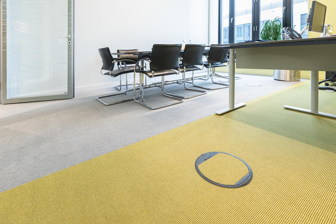 Ein heller Büroraum mit Arbeitstischen und Stühlen und gelbem Fußboden, in den ein diskretes Kabelelement eingelassen ist.