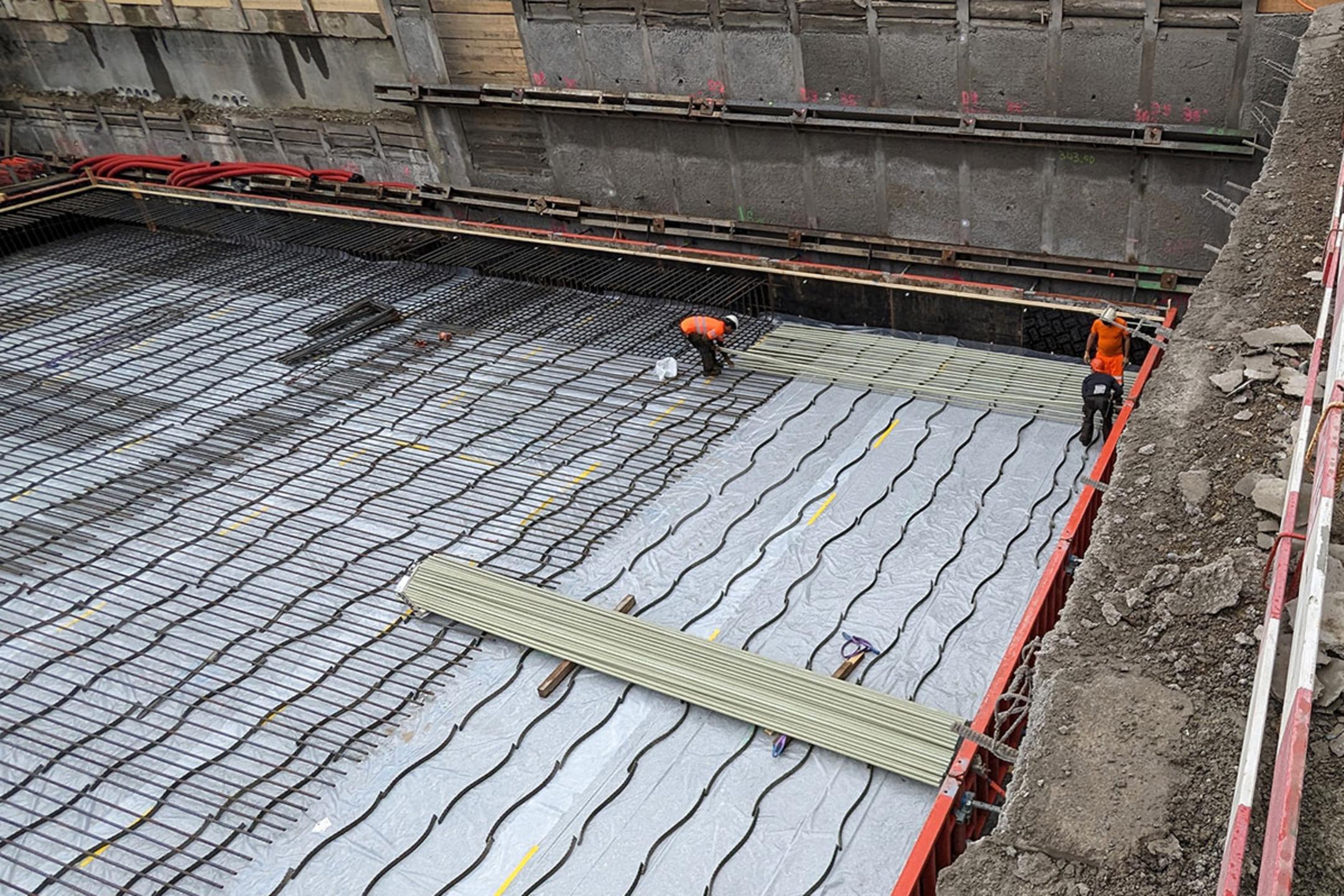 Bauarbeiter verlegen Bewehrung und Leitungen auf einer mit Folie abgedeckten Fundamentplatte in einer Baugrube