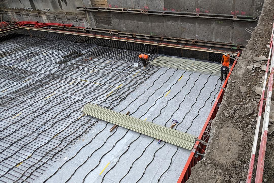 Bauarbeiter verlegen Bewehrung und Leitungen auf einer mit Folie abgedeckten Fundamentplatte in einer Baugrube