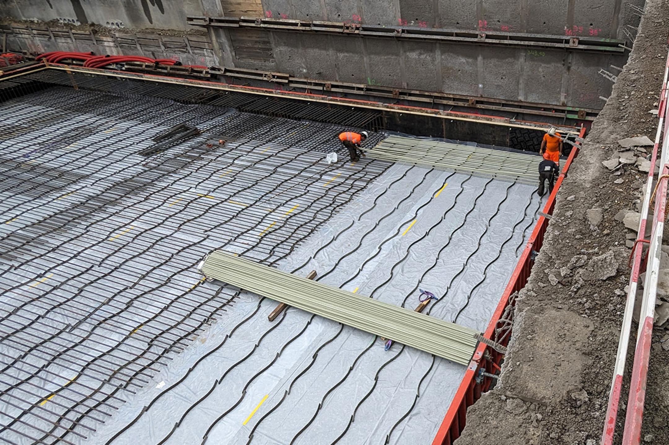 Bauarbeiter verlegen Bewehrung und Leitungen auf einer mit Folie abgedeckten Fundamentplatte in einer Baugrube