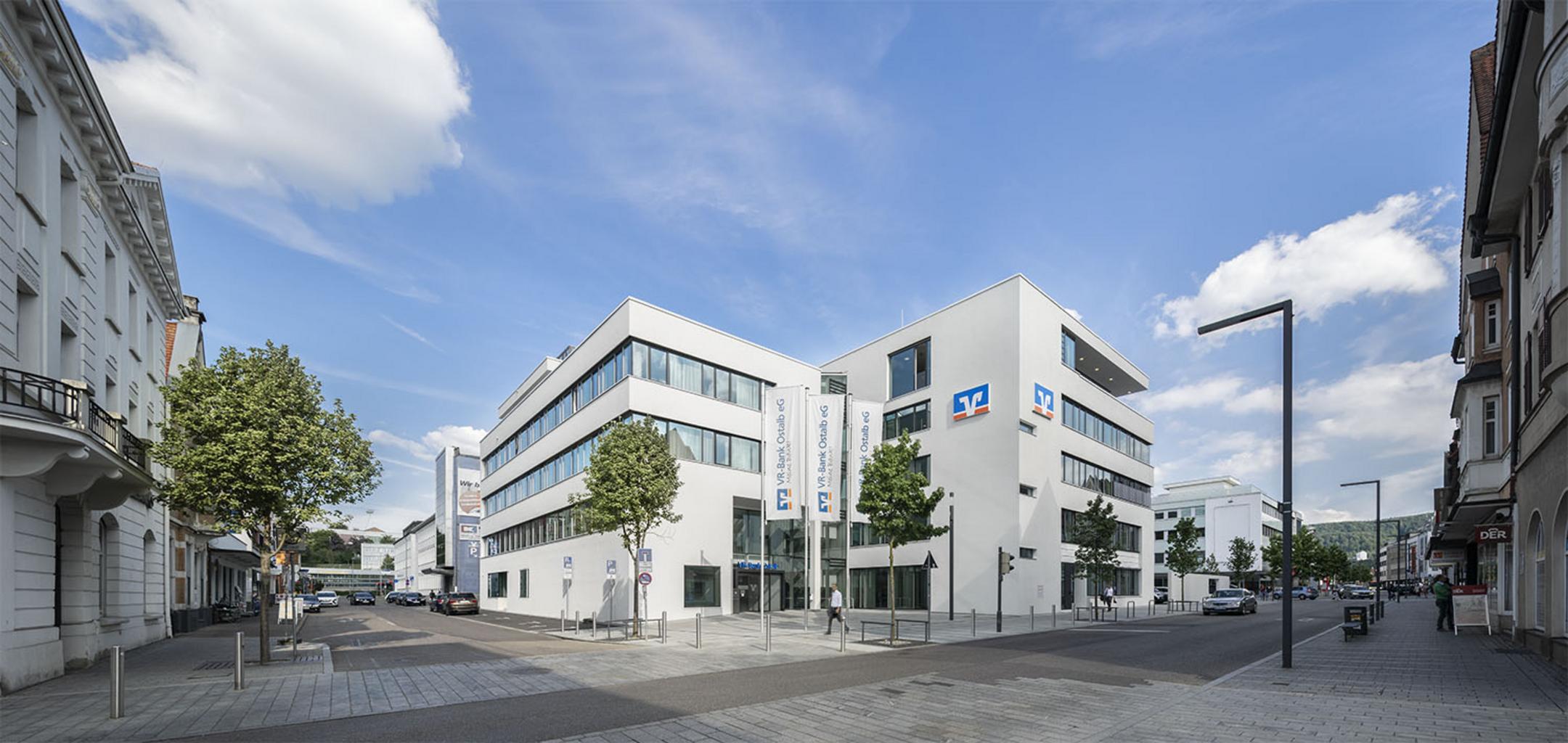 Frontalansicht eines weißen Gebäudes mit Volksbank-Logo mit großen Fenstern, Bäumen und geparkten Autos