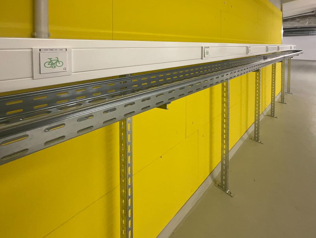 Metallschiene mit Fahrrad-Symbolen an gelber Wand, montiert auf Stahlträgern