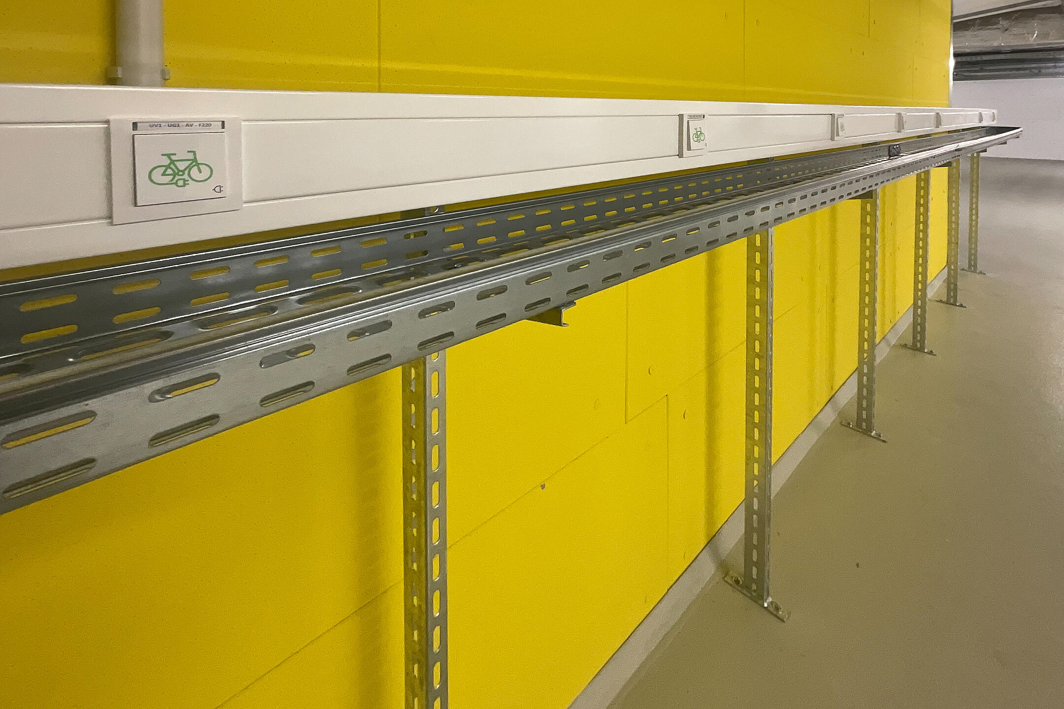 Metallschiene mit Fahrrad-Symbolen an gelber Wand, montiert auf Stahlträgern