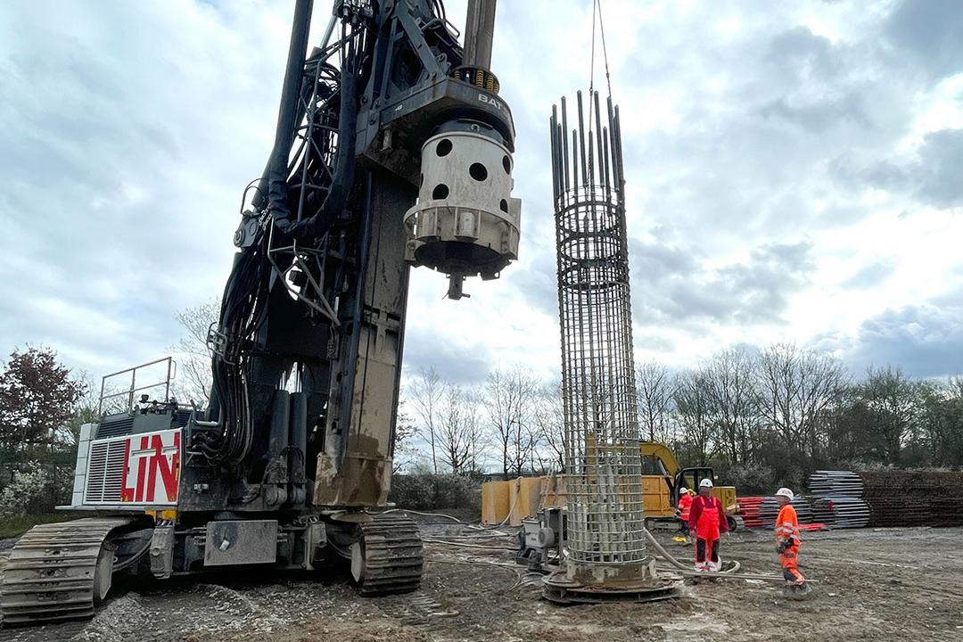 Baustelle mit großem Bohrgerät, Bewehrungskorb wird per Kran gehoben, mehrere Bauarbeiter in Warnkleidung beobachten