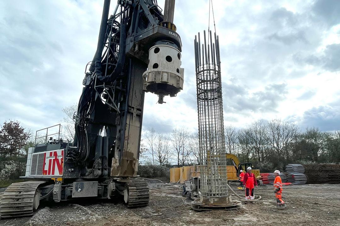 Baustelle mit großem Bohrgerät, Bewehrungskorb wird per Kran gehoben, mehrere Bauarbeiter in Warnkleidung beobachten