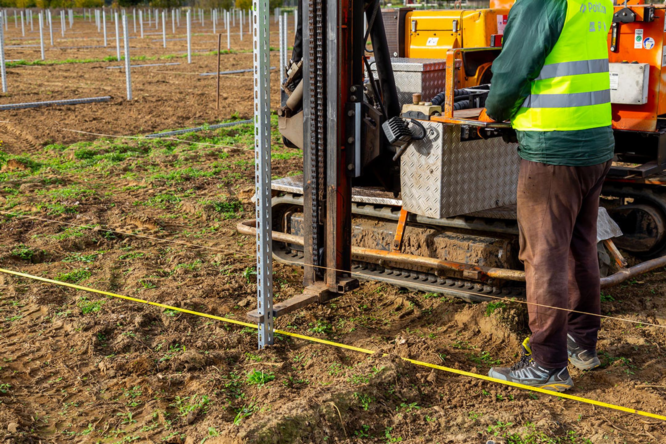 Arbeiter nutzt eine Maschine, um eine Stütze auf einem Solarfeld in den Boden zu rammen, dabei dienen ein gelbes Maßband und eine Schnur als Orientierung
