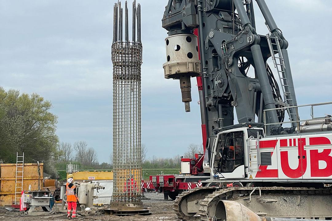 Bewehrungskorb wird in Boden gelassen, im Vordergrund ein Bohrgerät und Arbeiter, die den Vorgang bei bewölktem Himmel überwachen