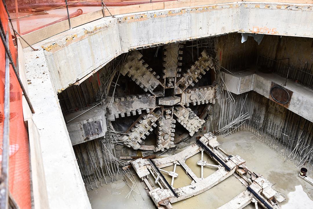 Tunnelbohrmaschine beim Vortrieb in einer Baugrube mit freiliegendem Schneidrad, Betonwänden und herausragender Bewehrung.