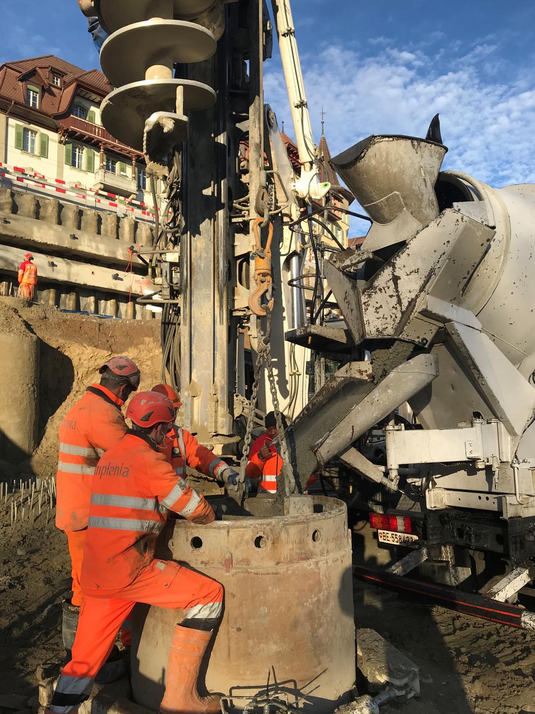 Arbeiter in Schutzkleidung gießen Beton aus LKW in zylindrische Schalung auf Baustelle mit Gebäuden im Hintergrund