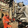 Arbeiter in Schutzkleidung gießen Beton aus LKW in zylindrische Schalung auf Baustelle mit Gebäuden im Hintergrund