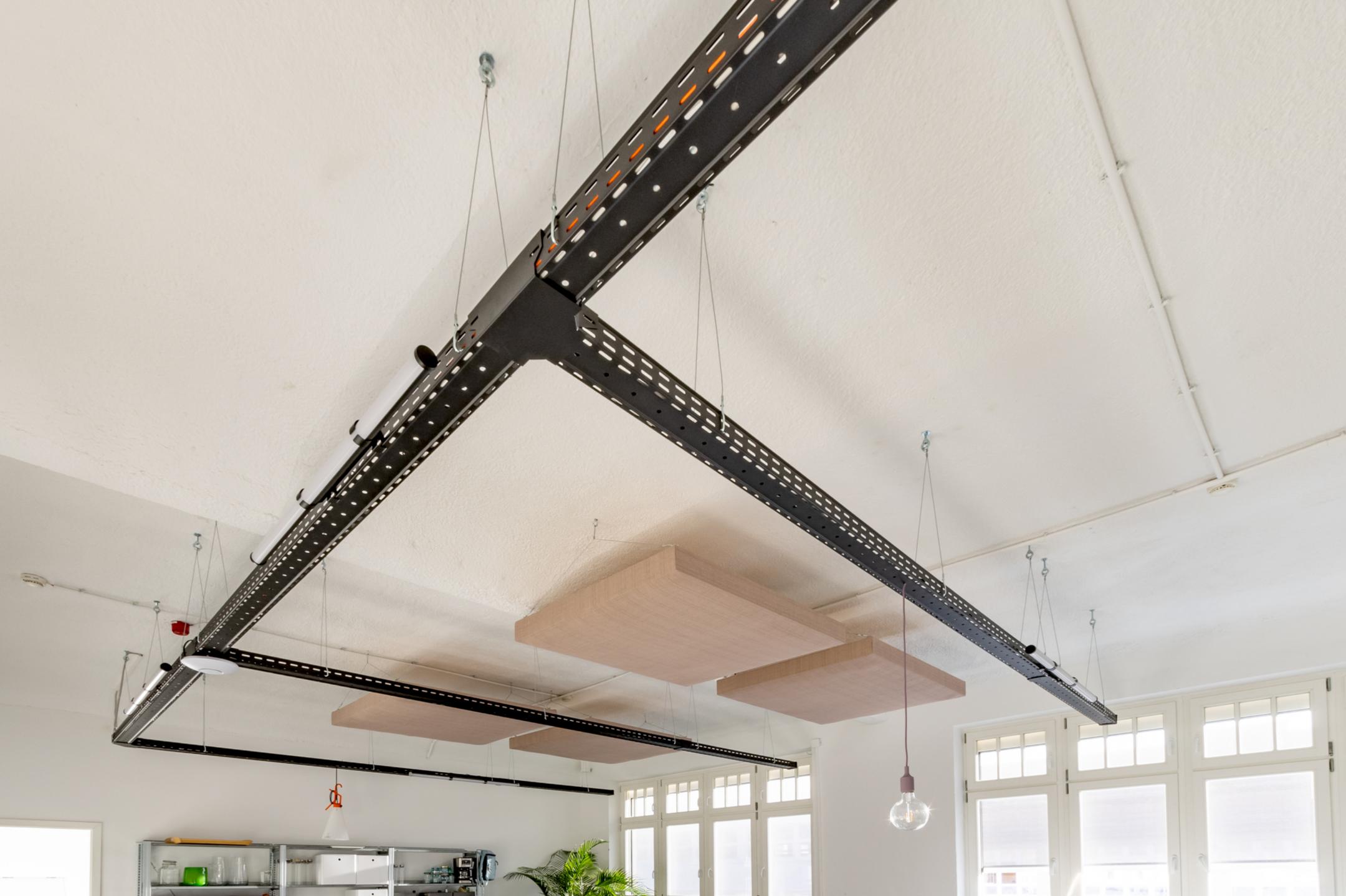 Schwarze Kabelrinnen mit T-Kreuzungen hängen an der Decke eines Raum, dazwischen sind Akustikelemente erkennbar.