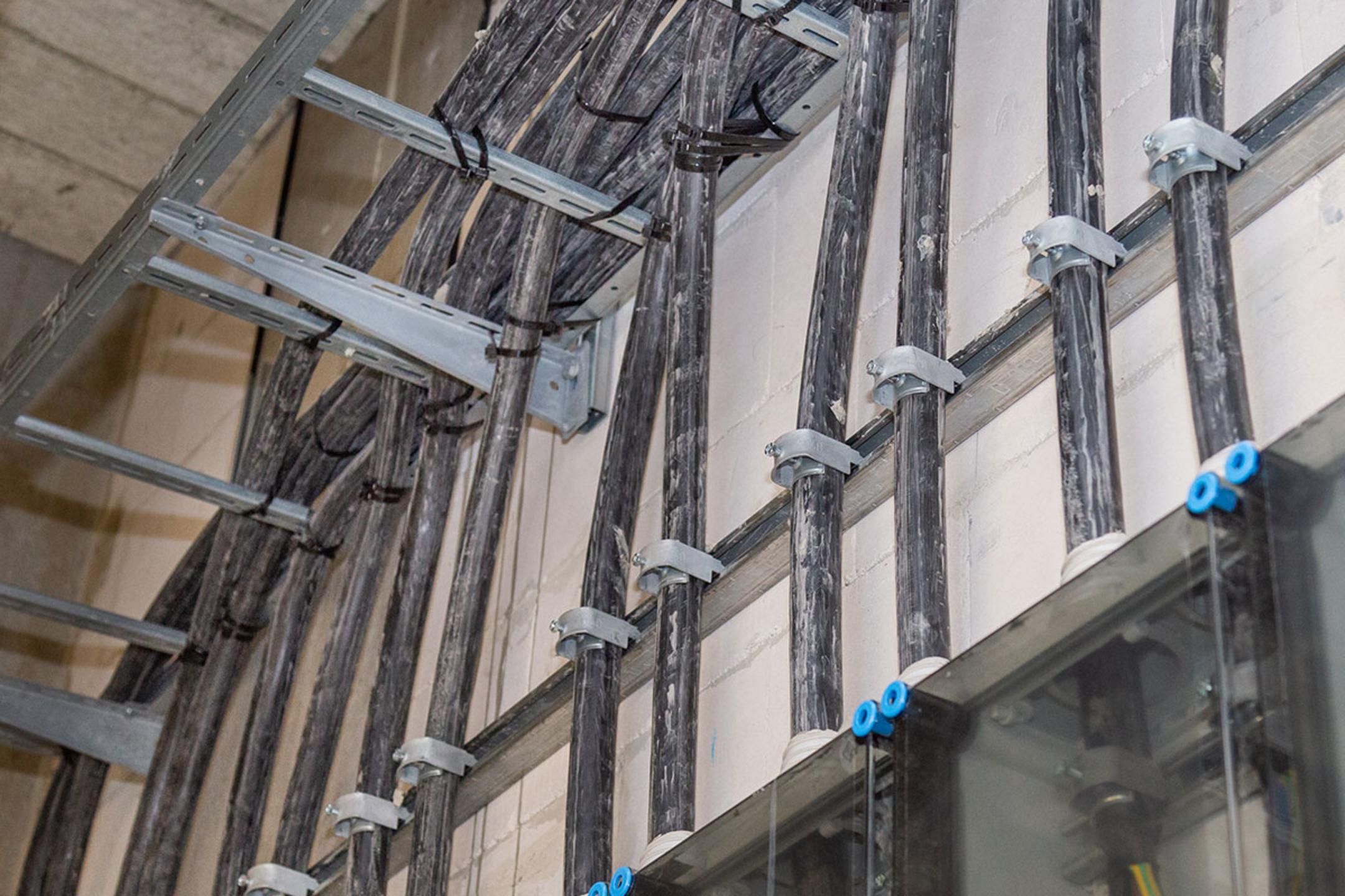Schwarze dicke Kabel, die mit Metallhalterungen an einer Wand entlanglaufen, mit Sicht auf Kabelleitern und Anschlusskästen