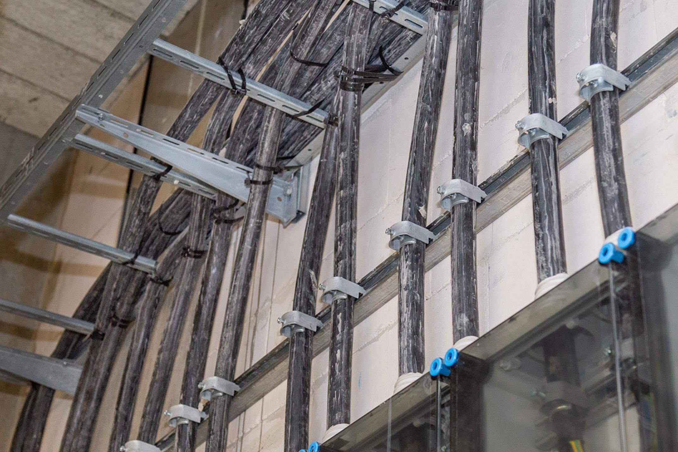 Schwarze dicke Kabel, die mit Metallhalterungen an einer Wand entlanglaufen, mit Sicht auf Kabelleitern und Anschlusskästen