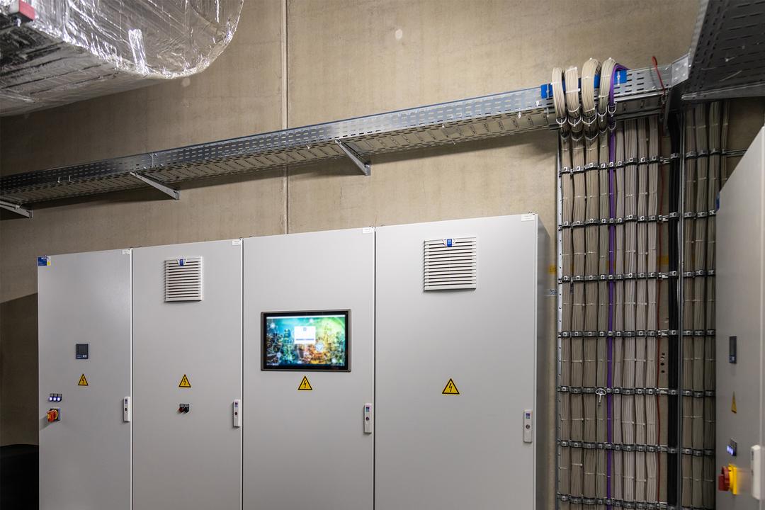 An Wand montierte Kabelrinne und Kabelleiter, bestückt mit mehrfarbigen Kabeln und befestigt mit Kabelschellen, Elektroschränke zentral.