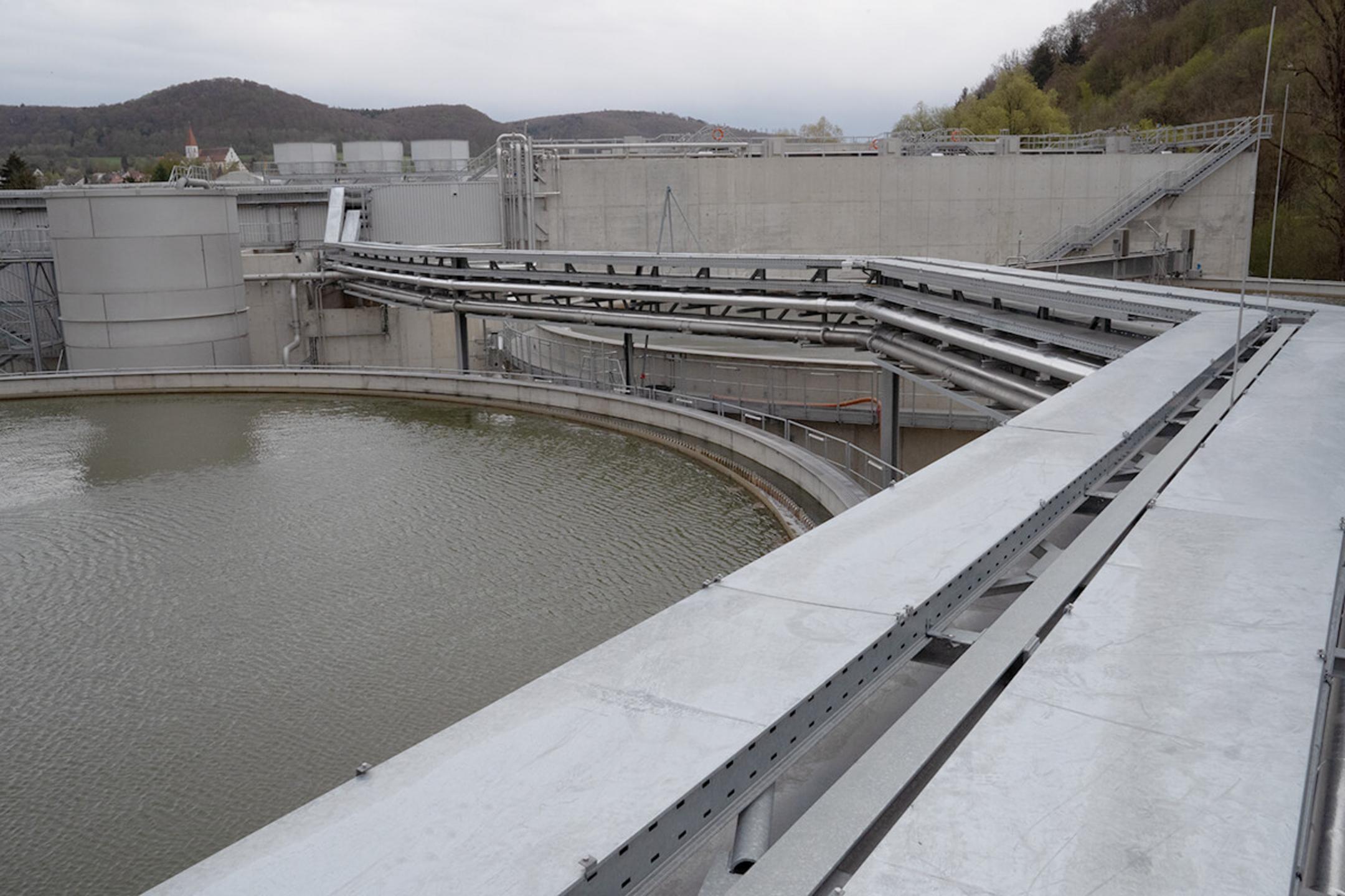 Blick auf ein rundes Wasserbecken auf einem Industriegelände mit Kabeltragsystemen an der Seite, bewölkter Himmel im Hintergrund