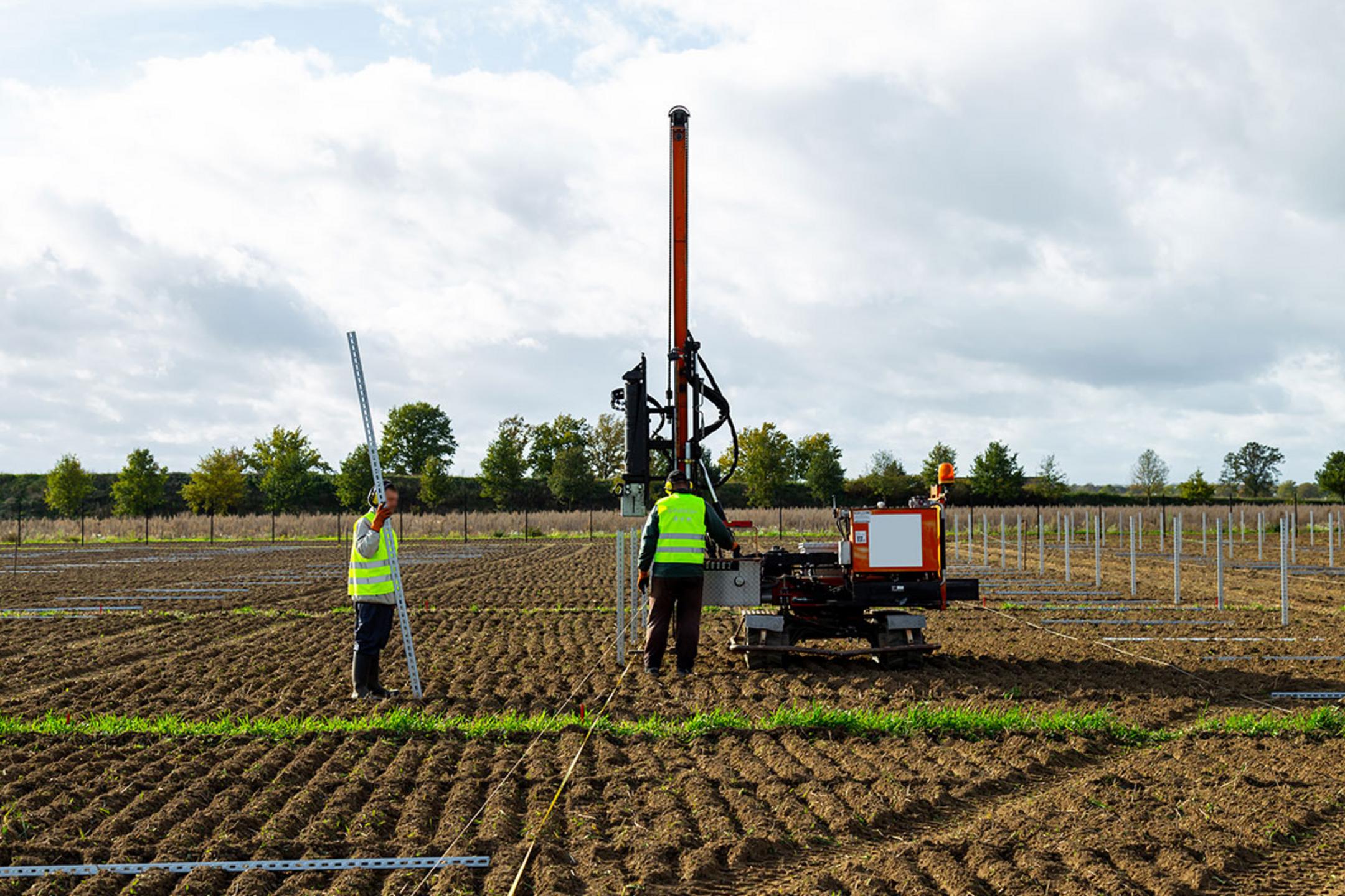 Zwei Arbeiter installieren Solarpanel-Stützen auf einem Feld, einer hält die Stütze, der andere bedient die Maschine