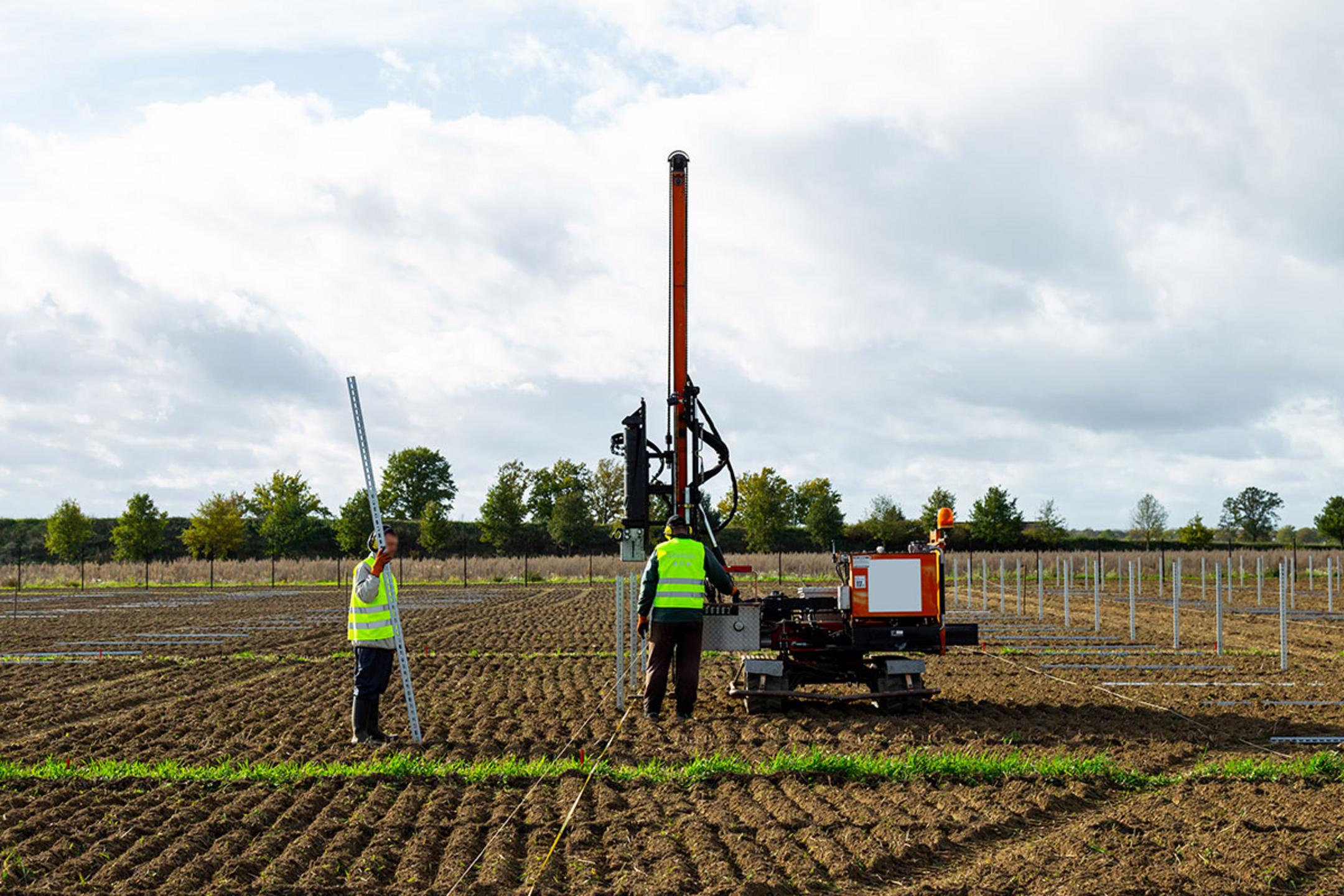 Zwei Arbeiter installieren Solarpanel-Stützen auf einem Feld, einer hält die Stütze, der andere bedient die Maschine