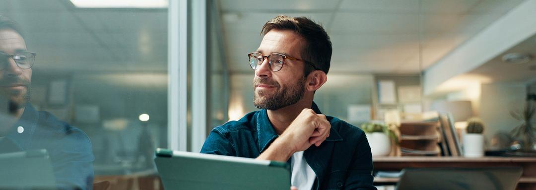 Ein Mann sitzt an einem Schreibtisch, hält ein Tablet in der Hand und guckt zufrieden aus dem Fenster.