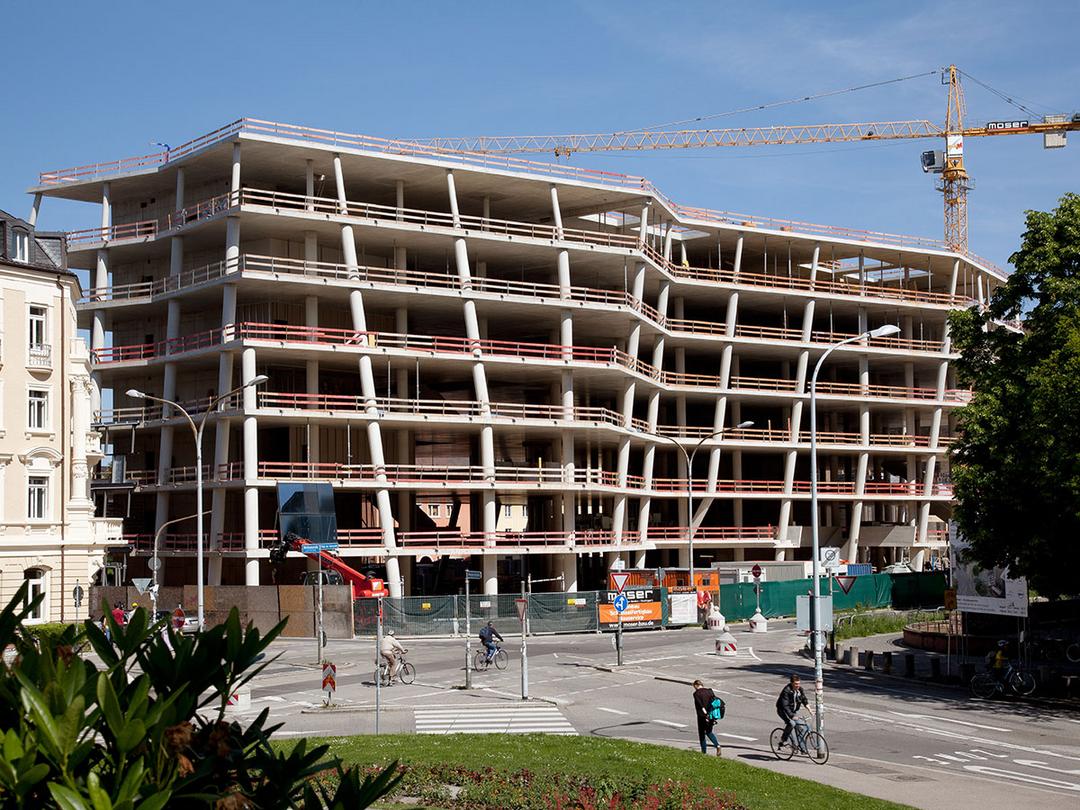Mehrstöckiger Rohbau mit Betonstützen und Baukran im Hintergrund, davor Straße mit Radfahrern und Fußgängern