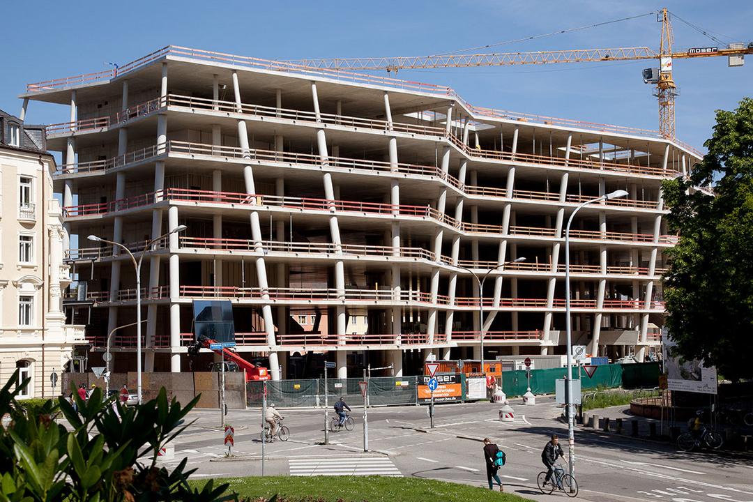 Mehrstöckiger Rohbau mit Betonstützen und Baukran im Hintergrund, davor Straße mit Radfahrern und Fußgängern