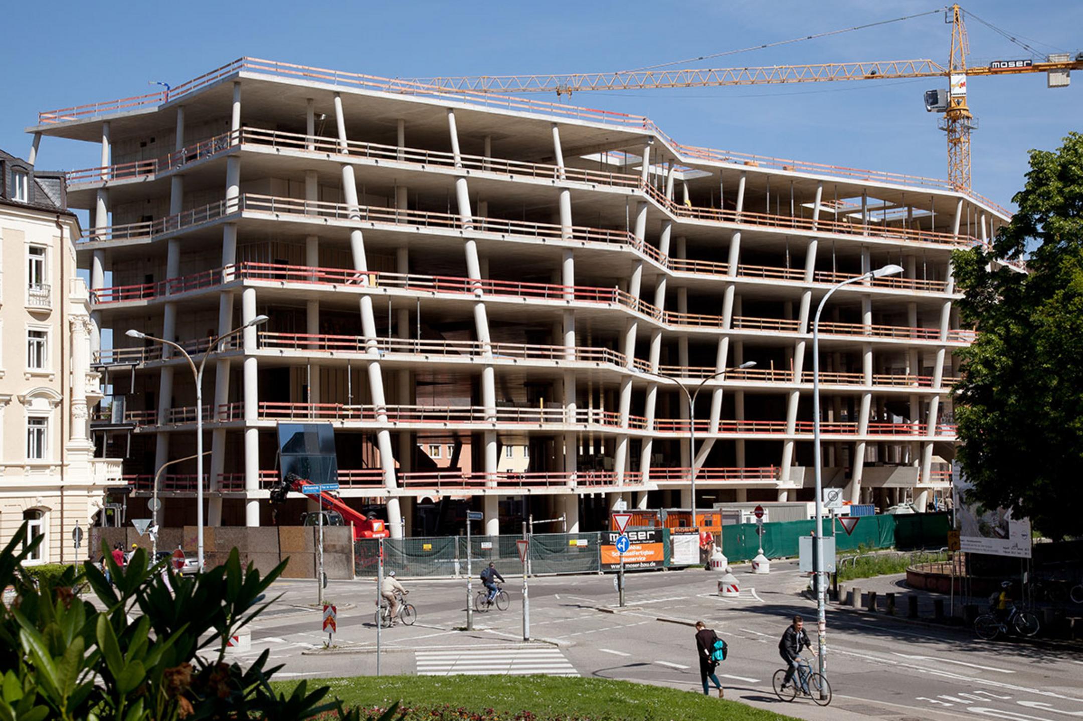 Mehrstöckiger Rohbau mit Betonstützen und Baukran im Hintergrund, davor Straße mit Radfahrern und Fußgängern