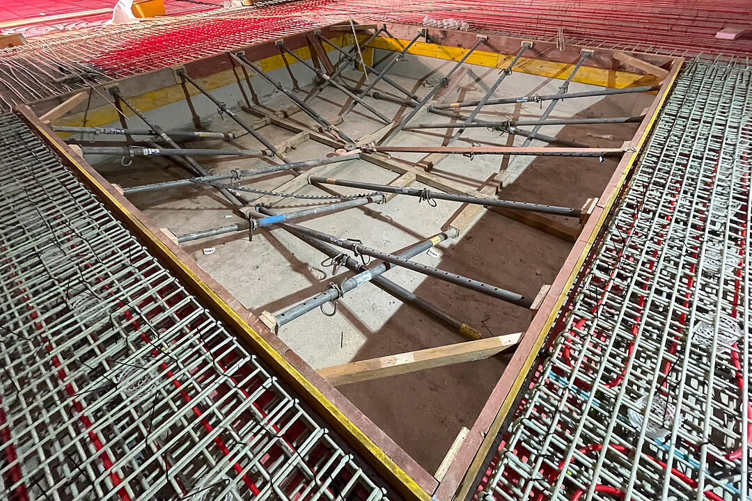 Baustellenaufnahme mit Bewehrungsmatten, roten Schläuchen auf dem Boden und ausgesteifter Schalung für eine Bodenaussparung vor dem Betonieren der Bodenplatte in einem Holzbau.