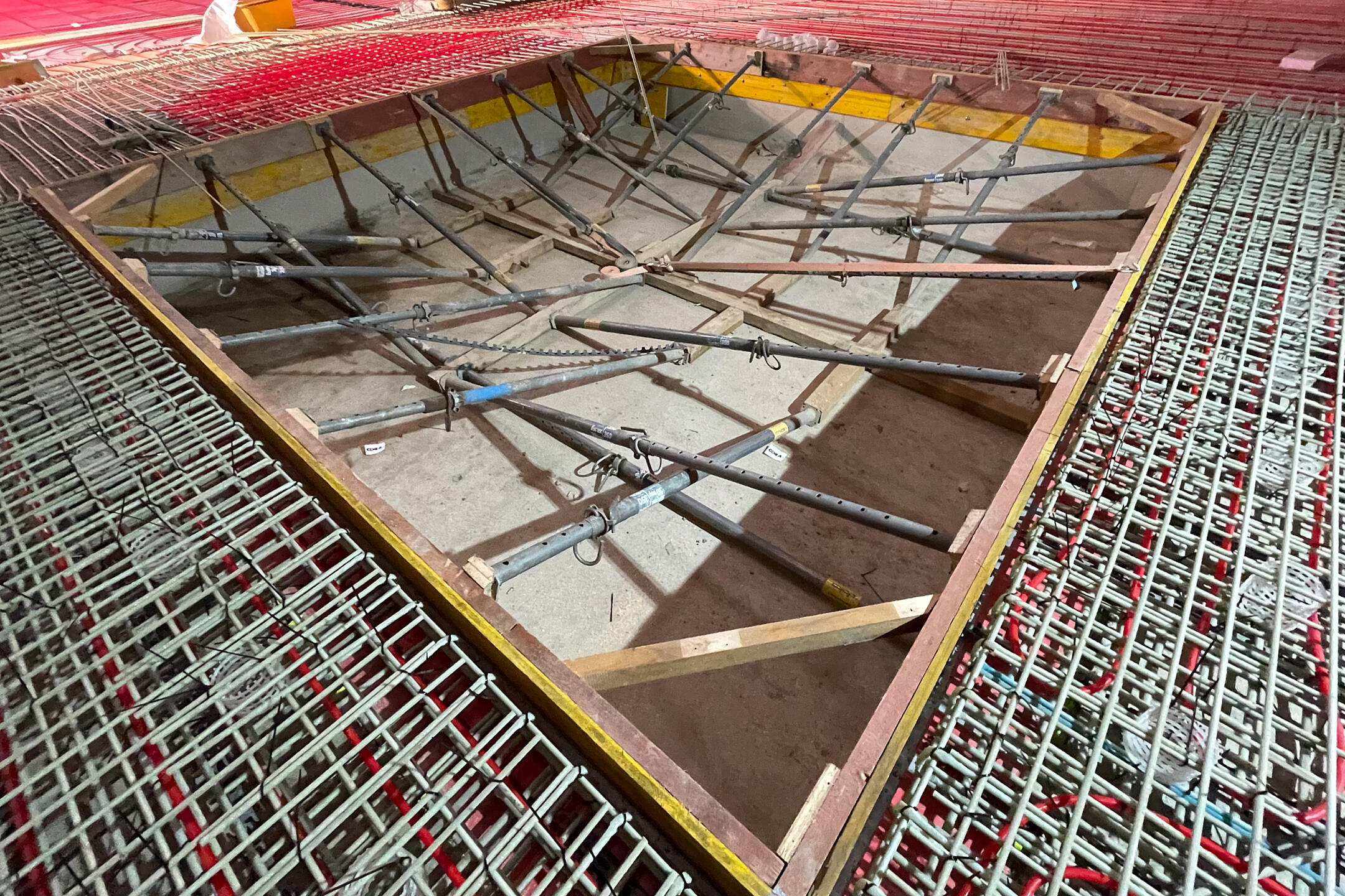 Baustellenaufnahme mit Bewehrungsmatten, roten Schläuchen auf dem Boden und ausgesteifter Schalung für eine Bodenaussparung vor dem Betonieren der Bodenplatte in einem Holzbau.
