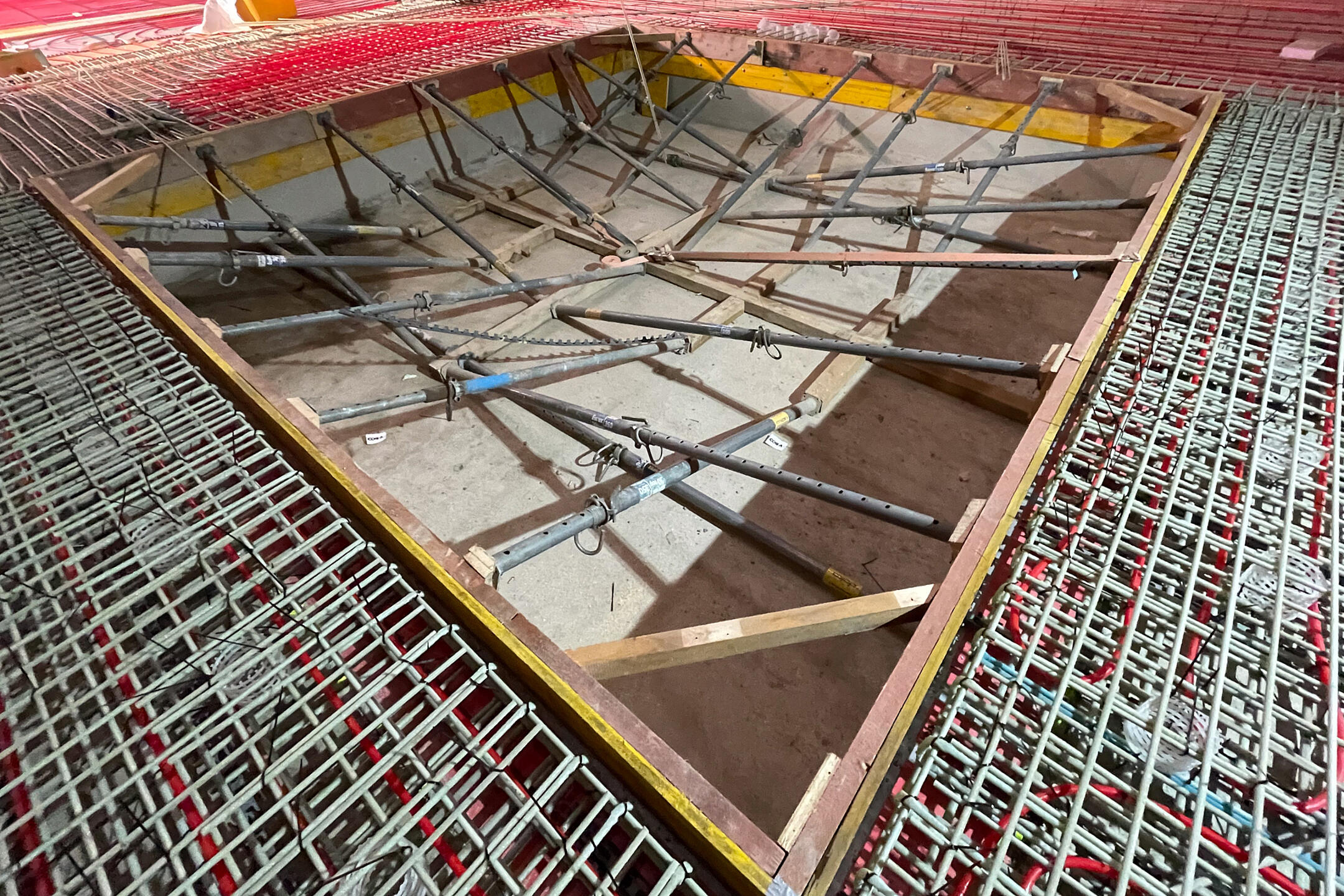 Baustellenaufnahme mit Bewehrungsmatten, roten Schläuchen auf dem Boden und ausgesteifter Schalung für eine Bodenaussparung vor dem Betonieren der Bodenplatte in einem Holzbau.