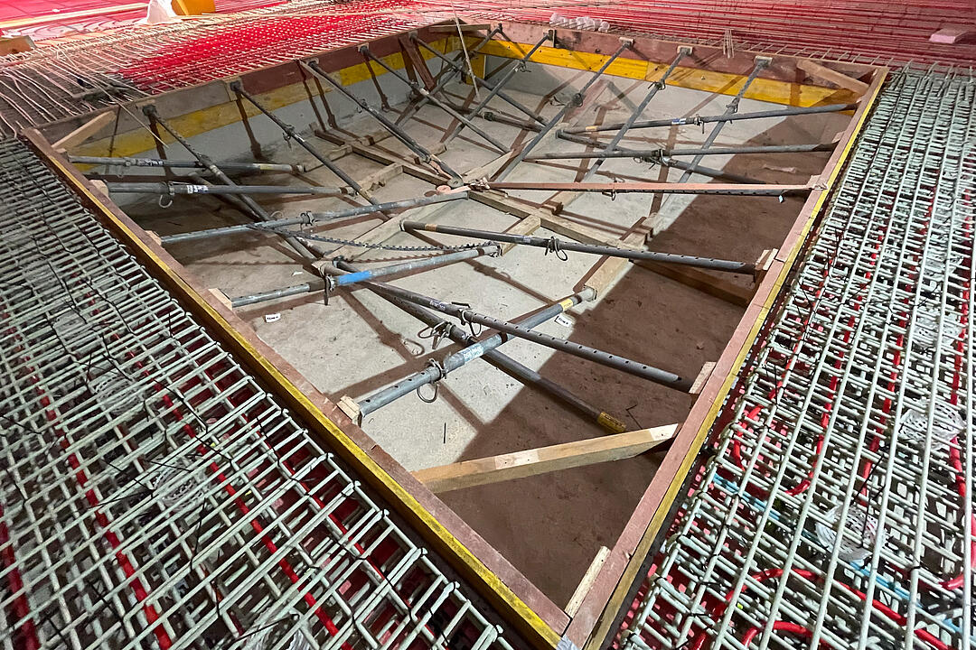 Baustellenaufnahme mit Bewehrungsmatten, roten Schläuchen auf dem Boden und ausgesteifter Schalung für eine Bodenaussparung vor dem Betonieren der Bodenplatte in einem Holzbau.