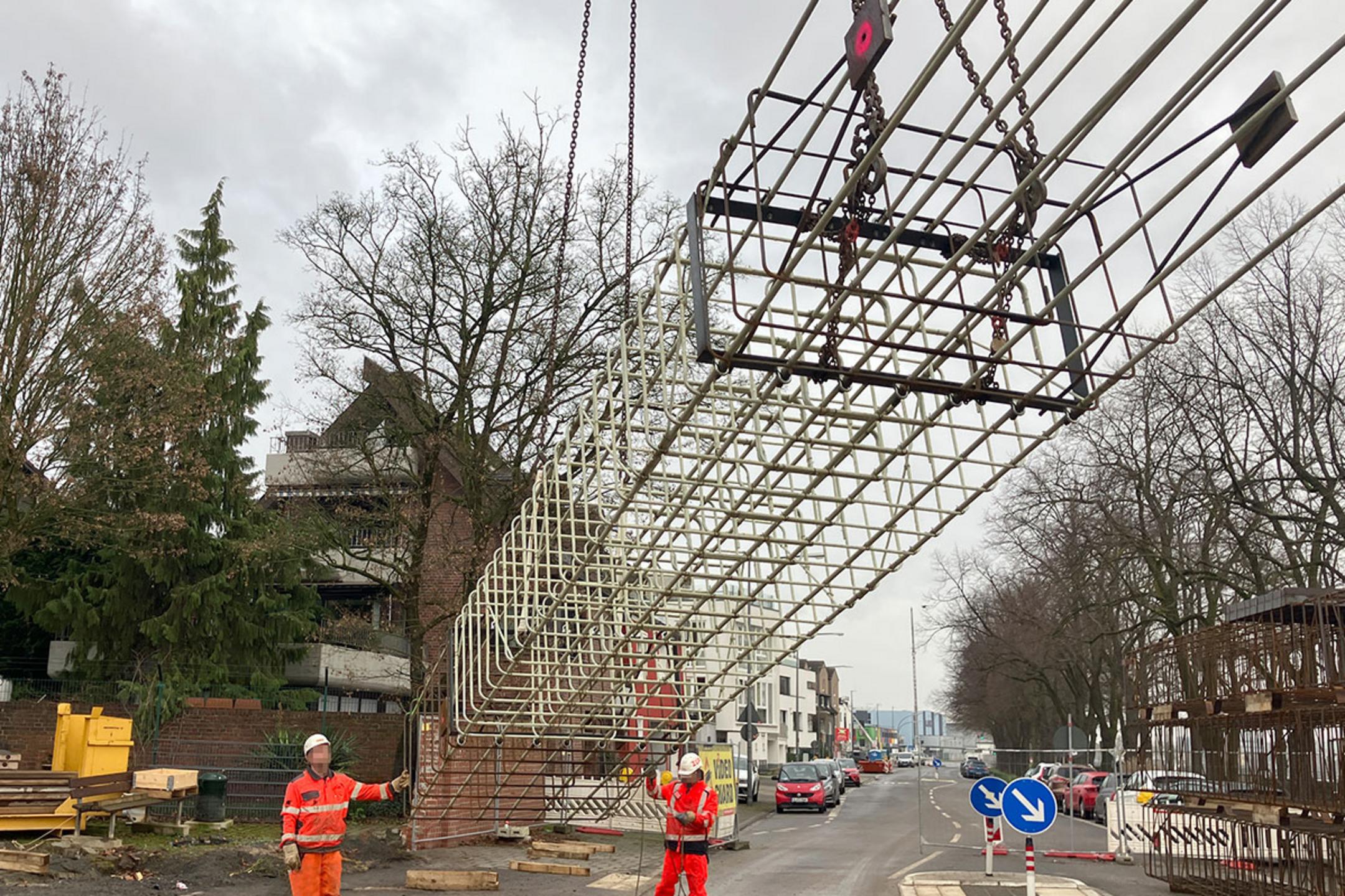 Baustelle an einer Straße, zwei Arbeiter in Warnkleidung steuern das Absetzen eines großen Bewehrungskorbs per Kran