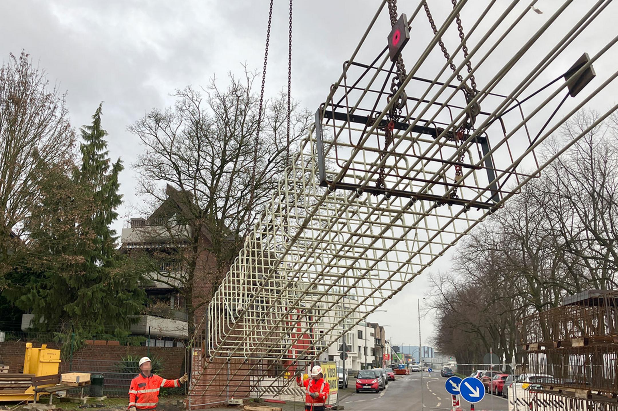 Baustelle an einer Straße, zwei Arbeiter in Warnkleidung steuern das Absetzen eines großen Bewehrungskorbs per Kran
