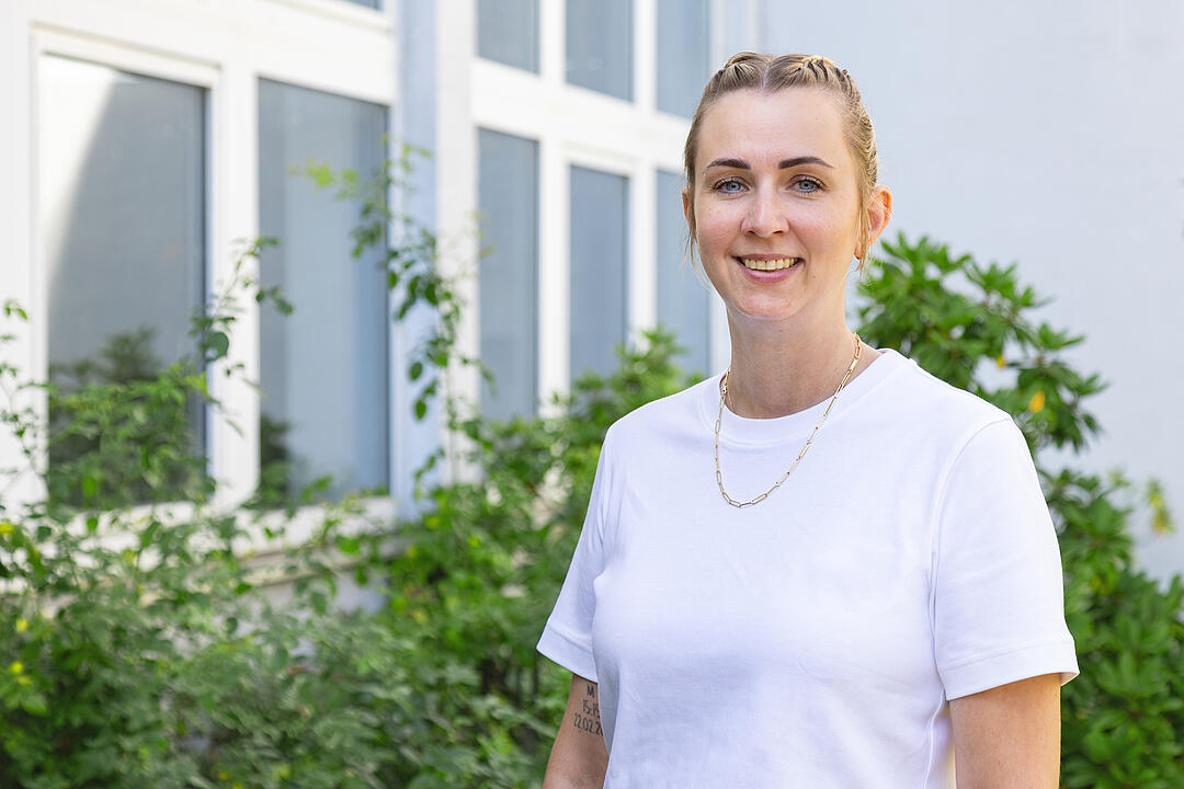 Portrait einer jungen Frau in einem weißen T-Shirt vor grünen Büschen und einer Fensterfront von einem Gebäude.