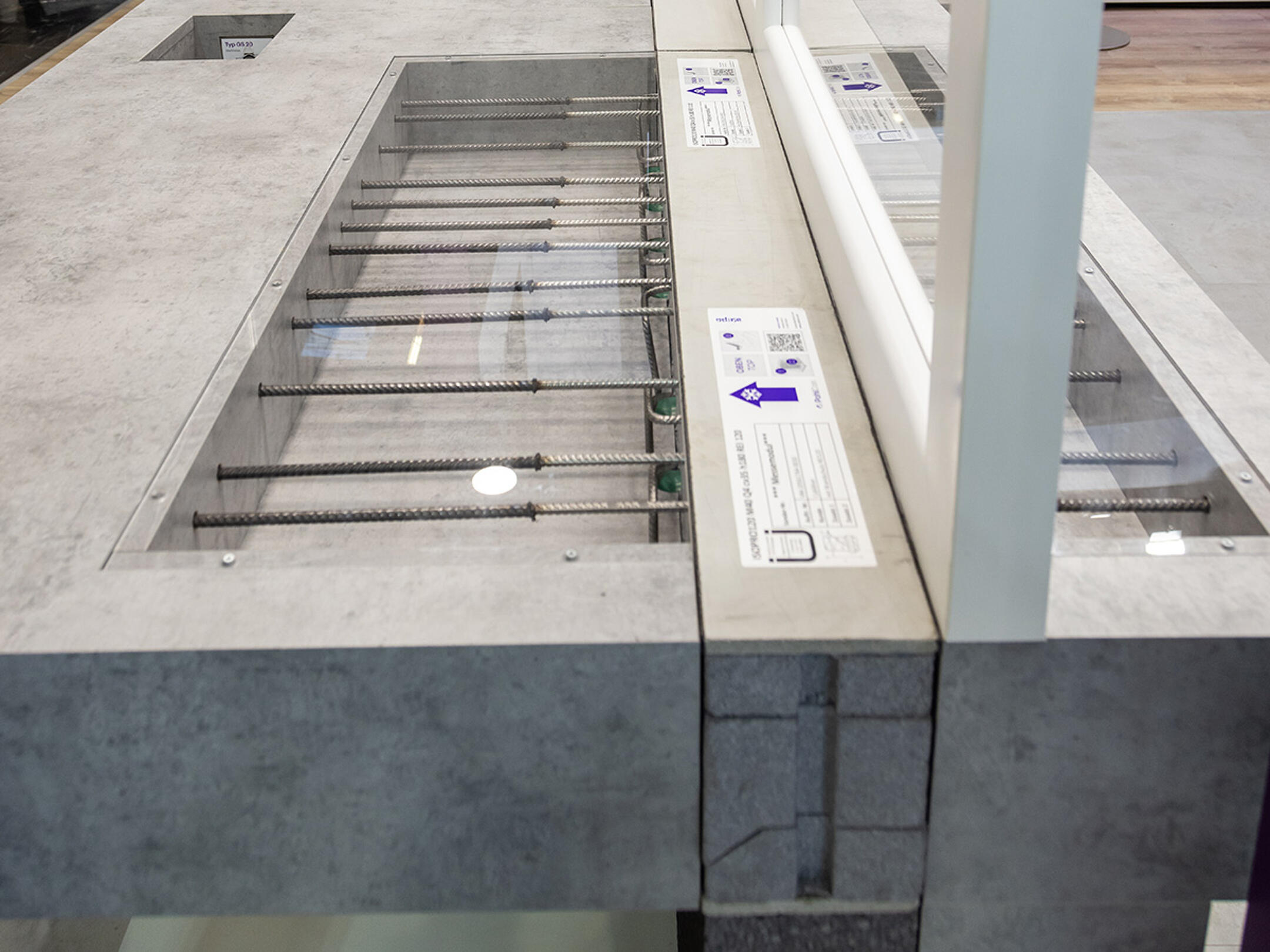 Betonbauteil als Ausstellungsmodell mit sichtbaren Bewehrungsstäben und integrierten Anschlussdetails unter Glas.