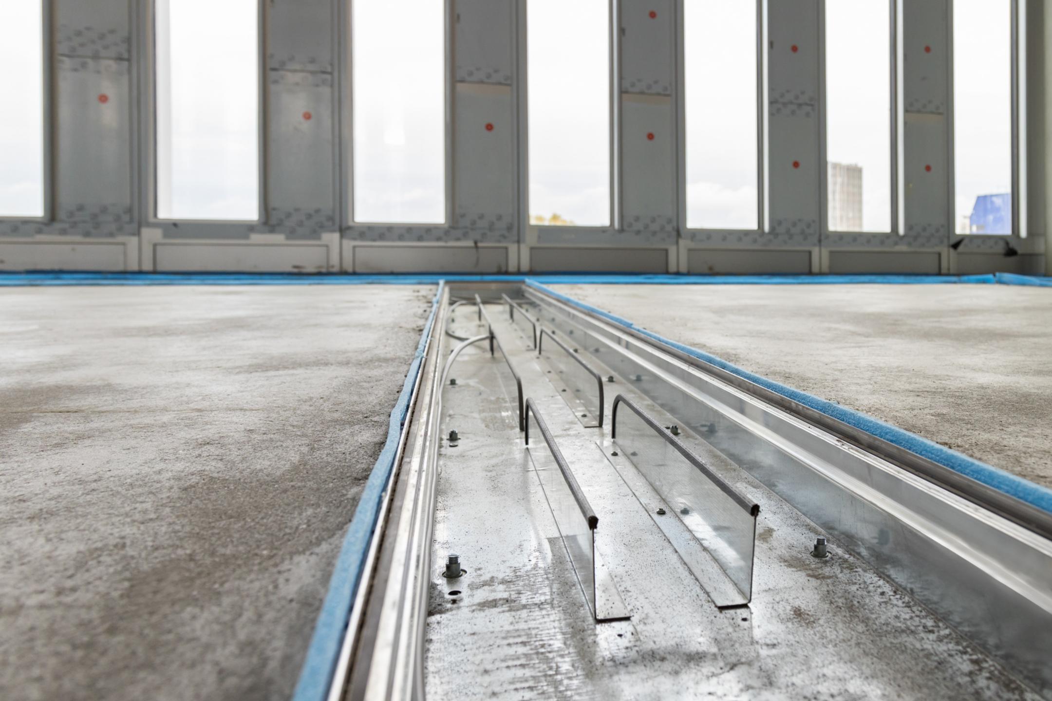 Geöffnetes Bodenkanalsystem im Estrichboden, im Hintergrund sind bodentiefe Fenster im Rohbau sichtbar