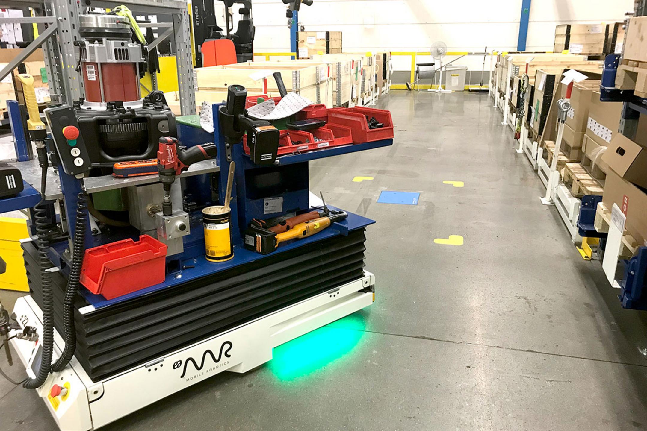 Automatisierter Transportroboter bewegt Waren durch ein Lager, im Hintergrund befindet sich eine blaue Ladeeinheit im Boden