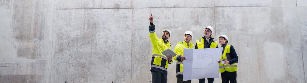Vier Personen mit Bauhelmen und Warnwesten betrachten auf einer Baustelle Baupläne, eine Person zeigt nach oben an die Wand.
