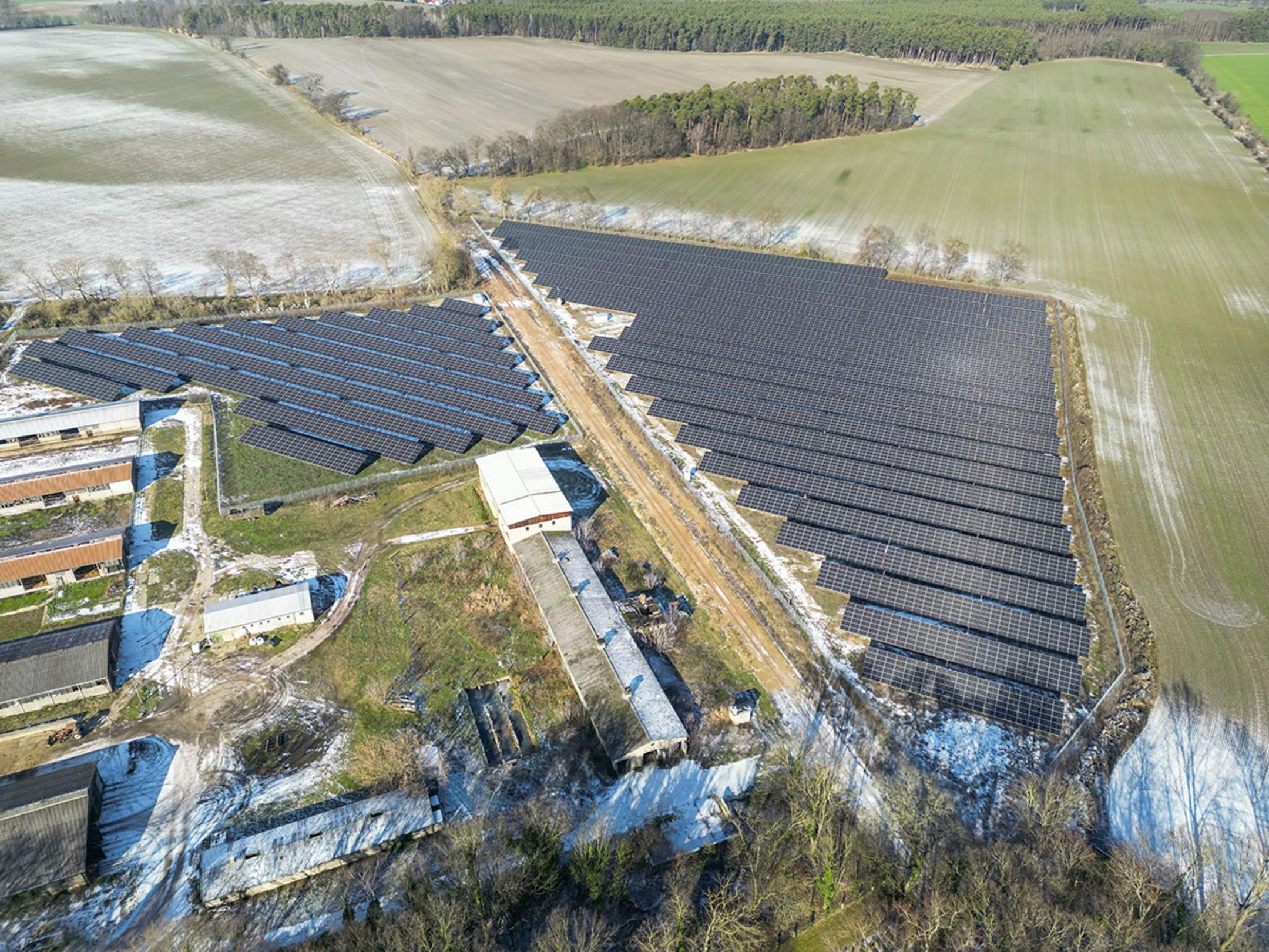 Luftaufnahme eines Solarparks mit angrenzenden Betriebsgebäuden, umgeben von Feldern, teilweise mit Schnee bedeckt