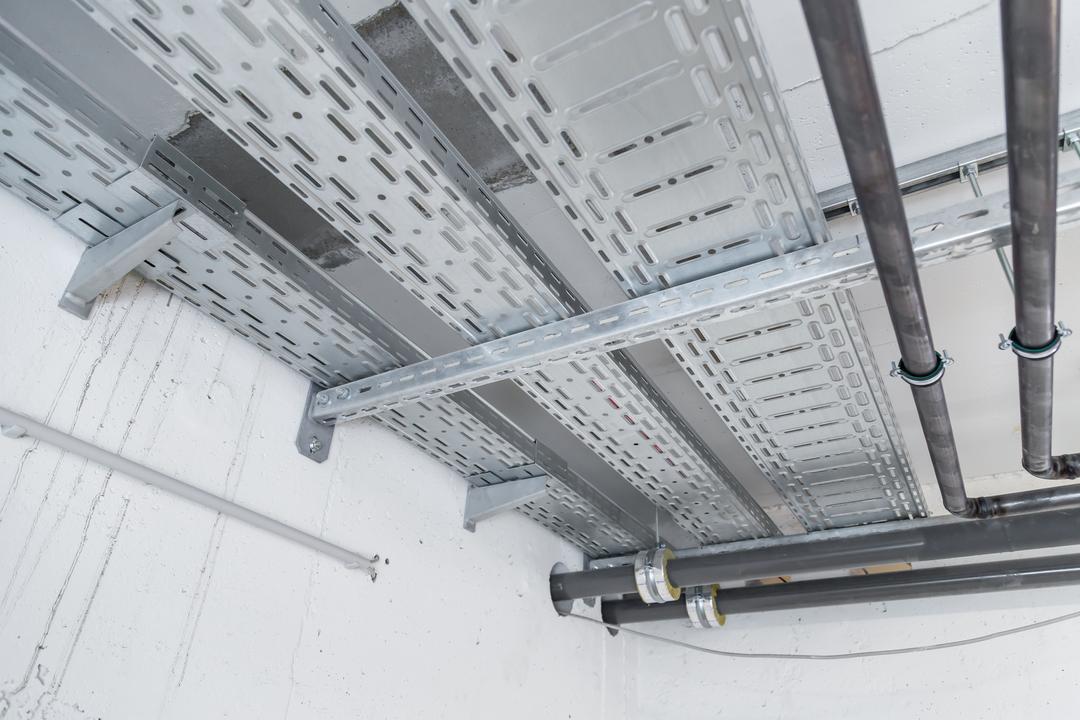 Industrieinstallation mit Kabeltrassen und Rohrleitungen an Decke und Wand in einem Technikraum mit Sichtbeton