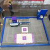 Ein Roboter vor einer Ladestation im Boden in einer Glasshowcase auf einer Messe, Besucher beobachten die Demonstration.