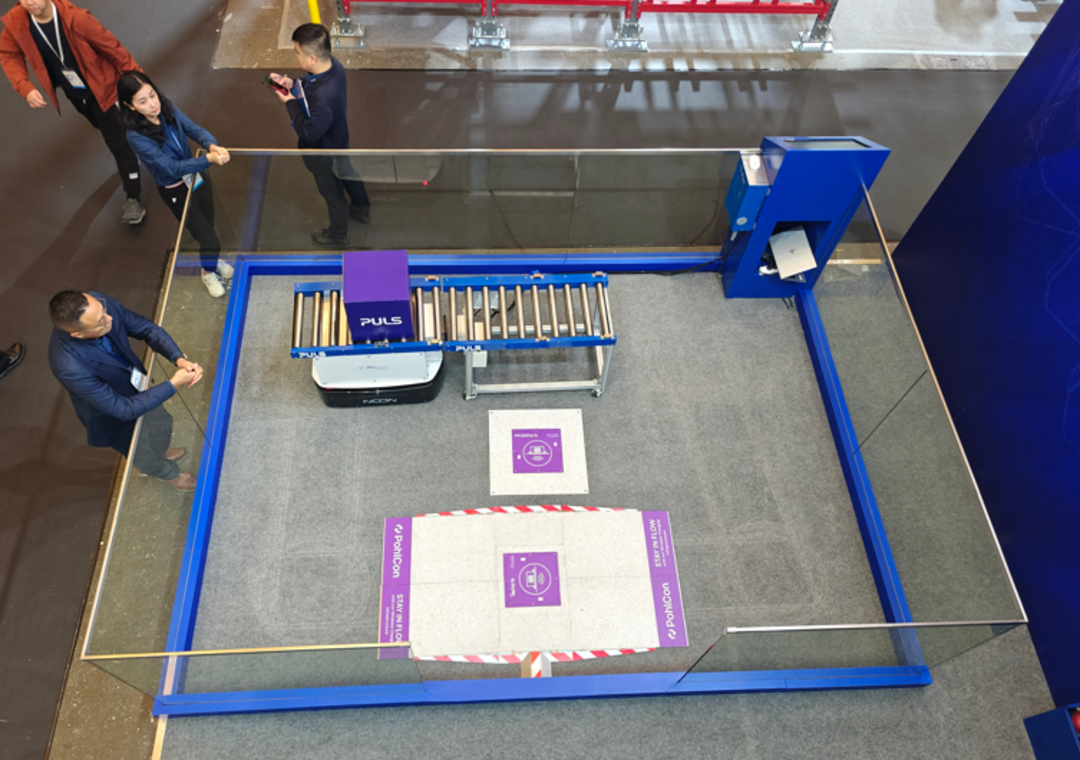 Ein Roboter vor einer Ladestation im Boden in einer Glasshowcase auf einer Messe, Besucher beobachten die Demonstration.